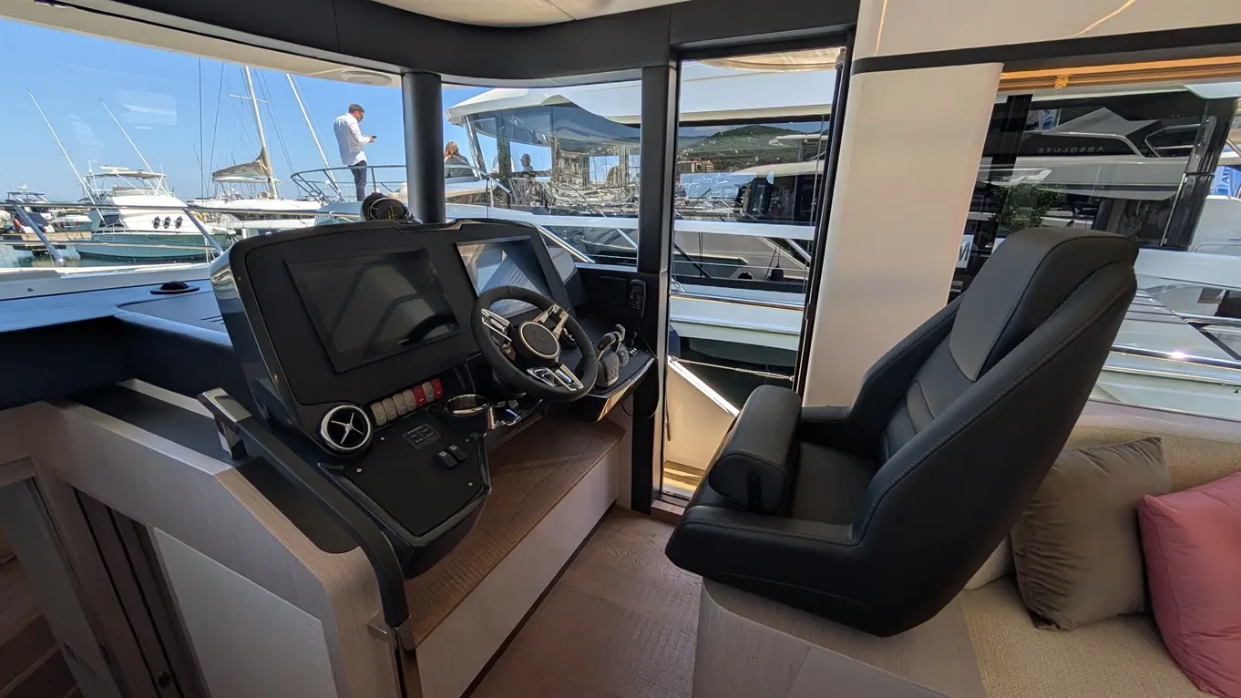 Luxury yacht helm with a steering wheel, electronic controls, and a comfortable seat, overlooking a marina with boats.
